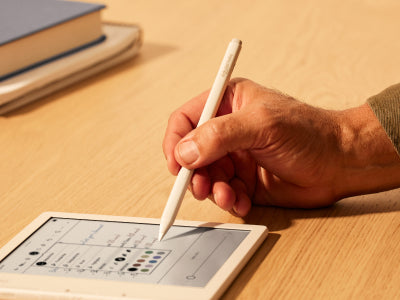 A hand is shown holding a Kobo Stylus 2 poised over a Kobo Libra Colour eReader on a flat surface. The tip of the stylus is about to select one of a range of pen colors shown in an options menu on the eReader screen.