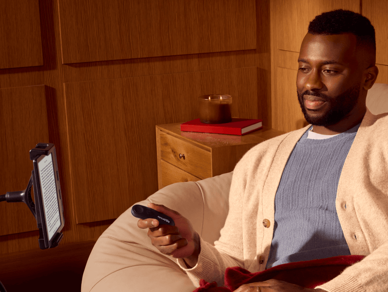 A man, seated comfortably, holds Kobo Remote in his right hand, while looking at a Kobo eReader on a bendable stand.