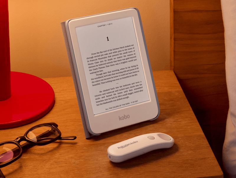 A white Kobo Clara Colour is propped up on table in its SleepCover with built-in stand, alongside a white Kobo Remote and a pair of eyeglasses.