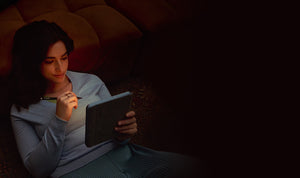 A woman reclining on a couch, lit by the soft light of a Kobo Libra Colour eReader she is holding, along with a Kobo Stylus 2.